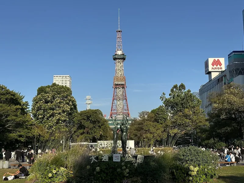 Sapporo TV Tower rising above green trees and flower beds in Odori Park under a clear blue sky.