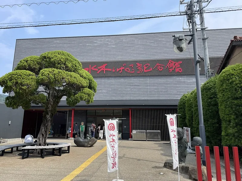 Mizuki Shigeru Museum in Sakaiminato under a blue summer sky, with sculpted trees and red signs.