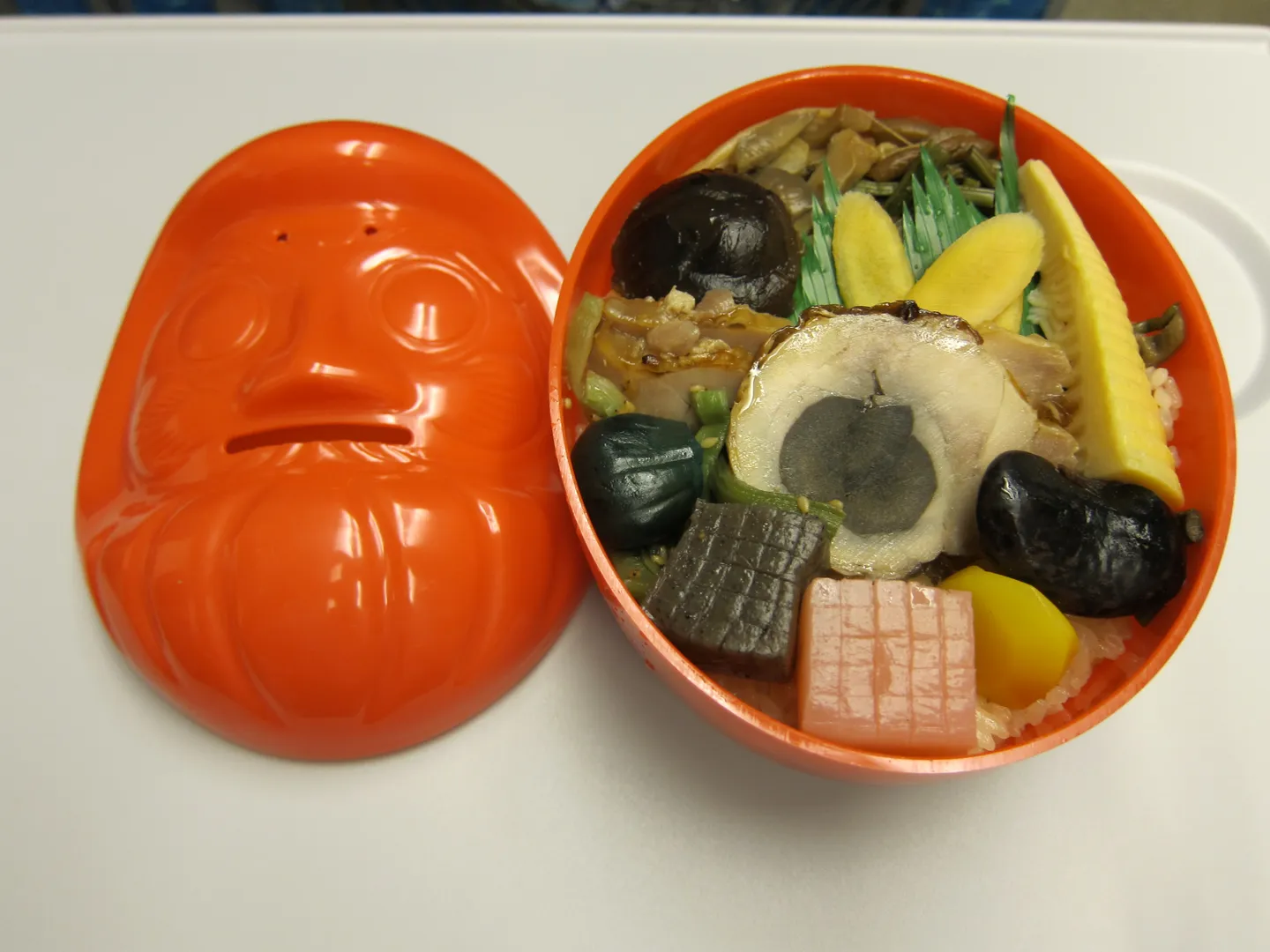 Egy daruma bento egy piros baba alakú edényben.