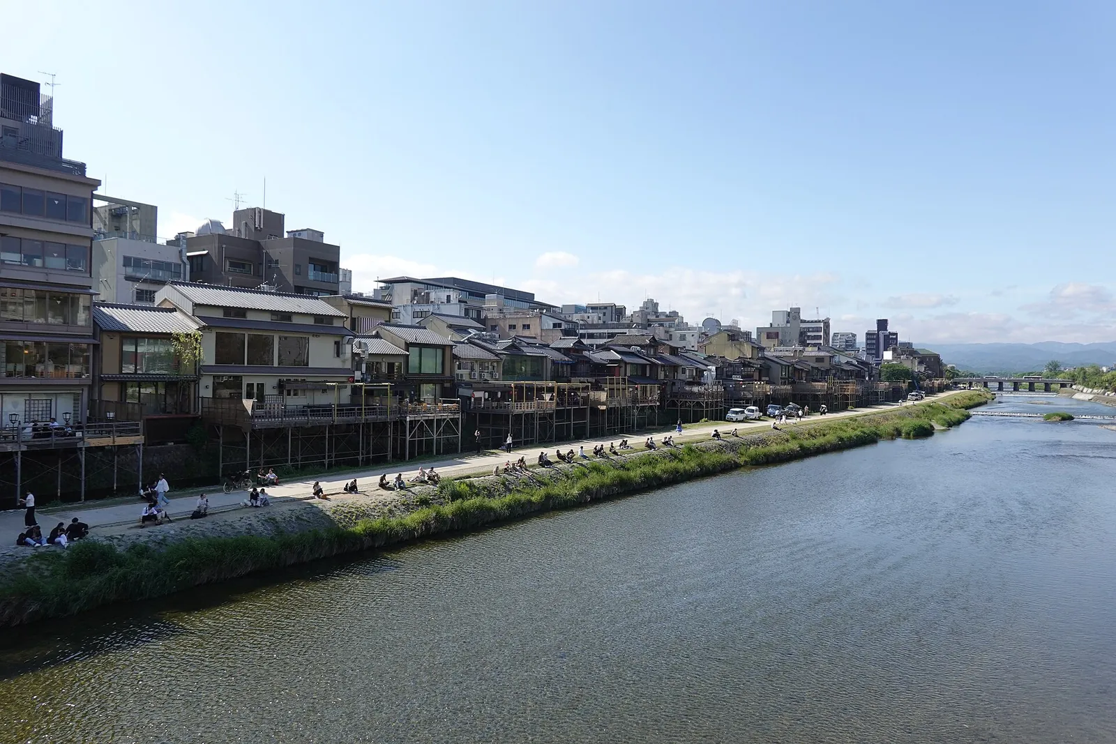 Stadthäuser und Restaurants mit hölzernen Terrassen am Fluss entlang des Westufers des Kamo im Zentrum von Kyoto, mit Menschen, die unter klarem blauem Himmel am Damm sitzen