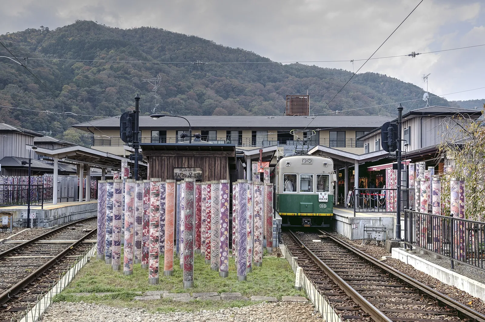 Eine grüne Randen-Straßenbahn am Bahnhof Arashiyama, mit den stoffumwickelten Pfosten des Kimono Forest neben den Gleisen und bewaldeten Hügeln dahinter