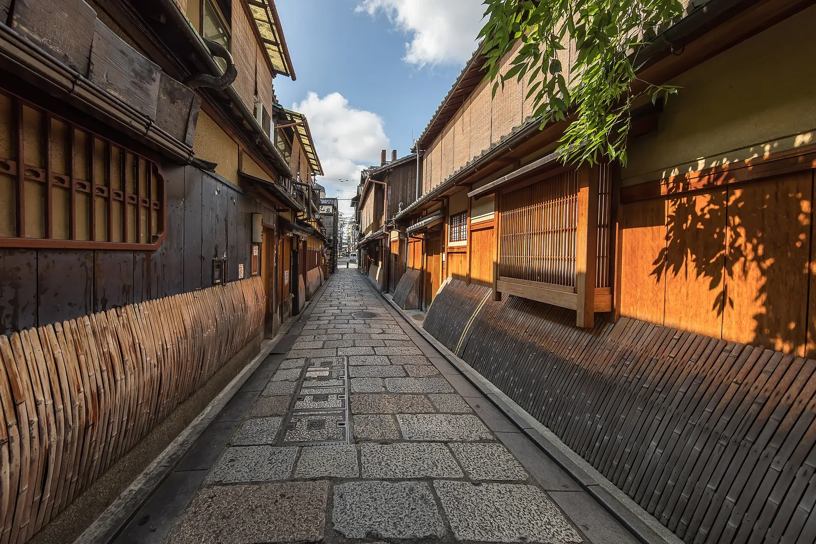 Eine schmale, steingepflasterte Gasse in Gion, gesäumt von dunklen Holzhäusern, Lattenfenstern und geschwungenen Bambuszäunen, im Licht der späten Nachmittagssonne