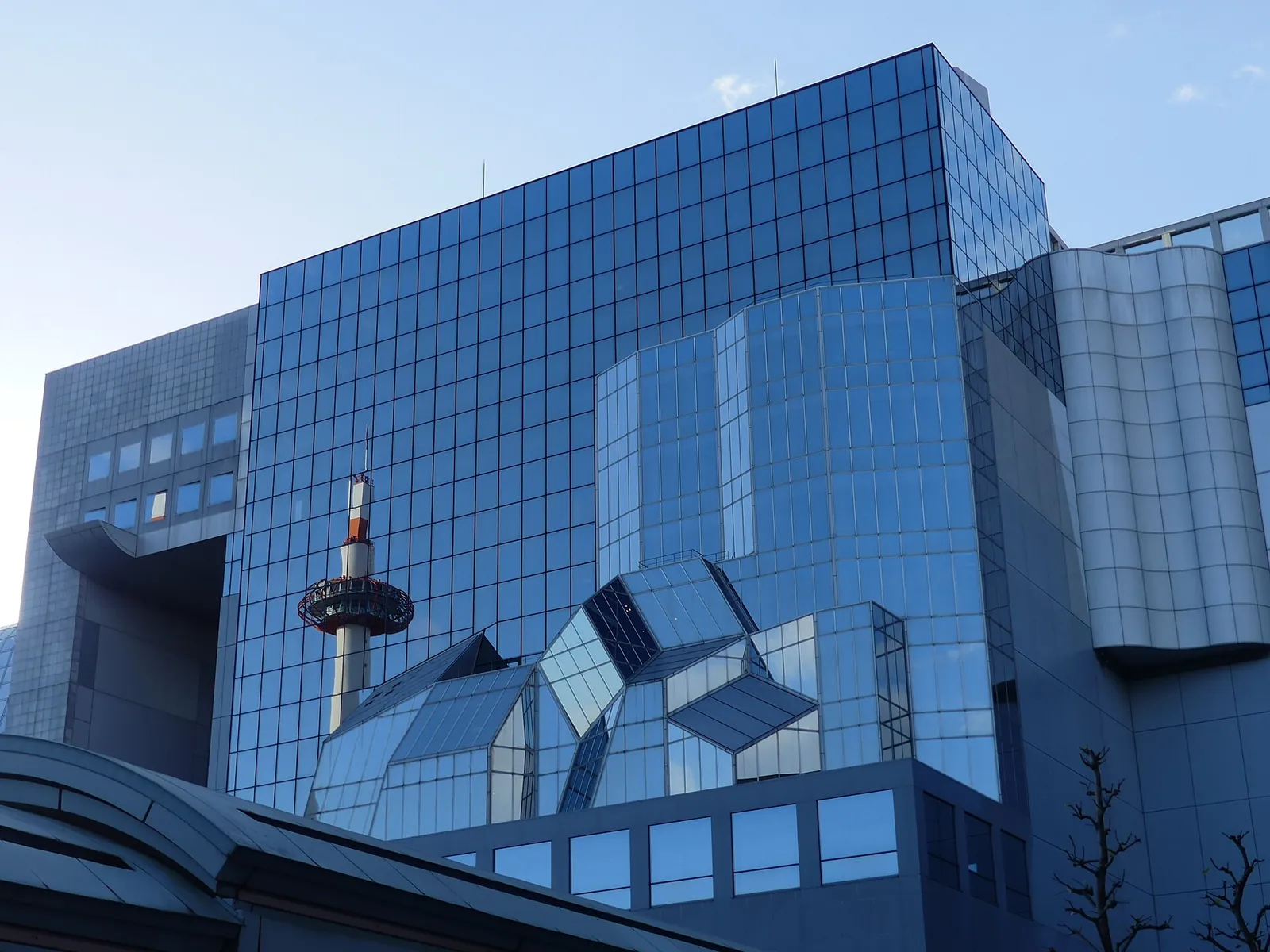 Die Glasfassade des Hauptgebäudes des Bahnhofs Kyoto, in deren Scheiben sich der Kyoto Tower unter blassblauem Himmel spiegelt