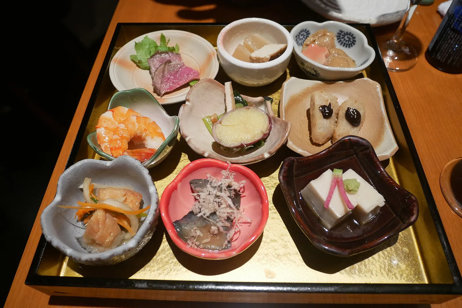 Neun kleine Gerichte der Kyoto-Hausmannskost auf einem goldenen Tablett, darunter Garnelen, geschmortes Gemüse, geschnittenes Rindfleisch, Makrele mit Bonitoflocken und ein Quadrat Tofu