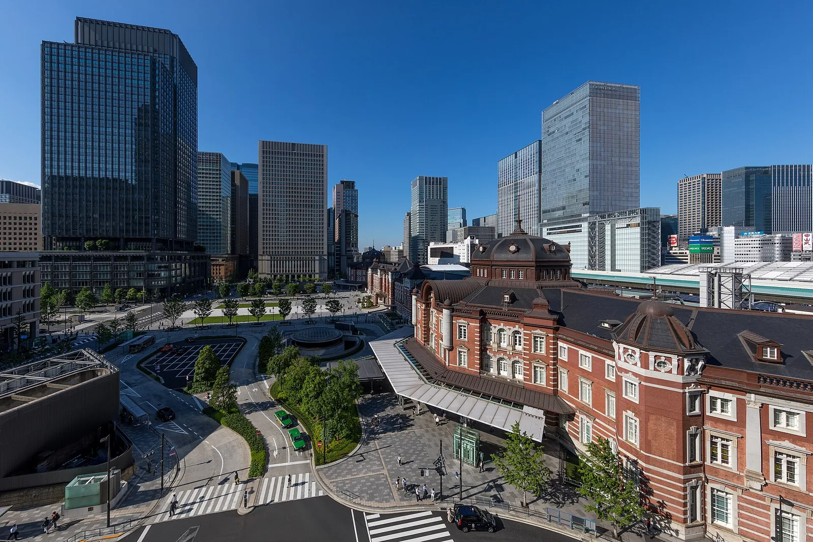 L'edificio Marunouchi in mattoni rossi della stazione di Tokyo accanto a una piazza alberata, con le torri di vetro di Marunouchi sullo sfondo sotto un cielo azzurro