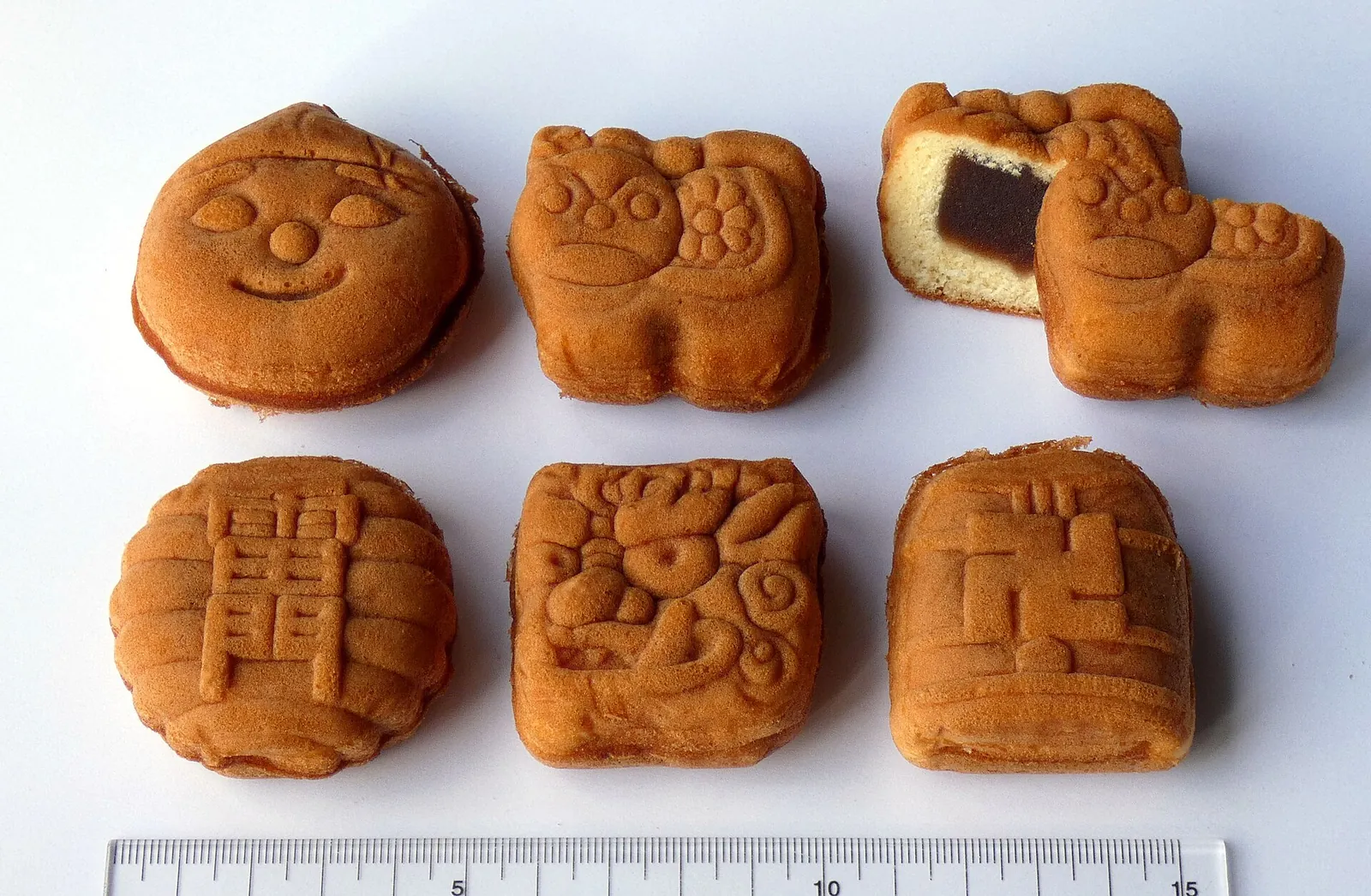 Sei dolci ningyo-yaki dorati accanto a un righello, con impresso un viso sorridente e altre figure, uno spezzato a metà per mostrare la pasta dolce di fagioli