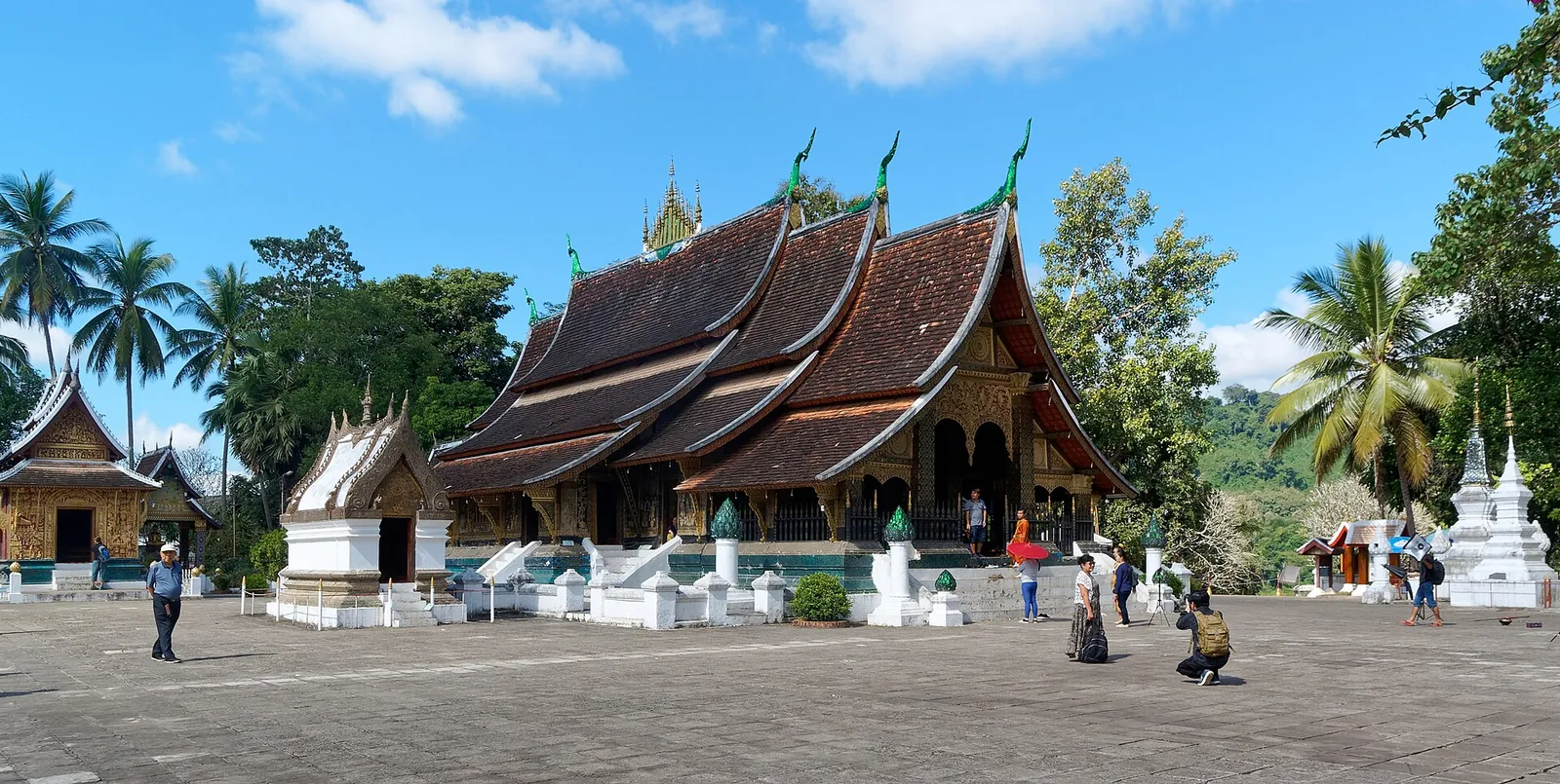 Splývavé chrámové střechy a dlážděné nádvoří ve Wat Xieng Thong v Luang Prabangu
