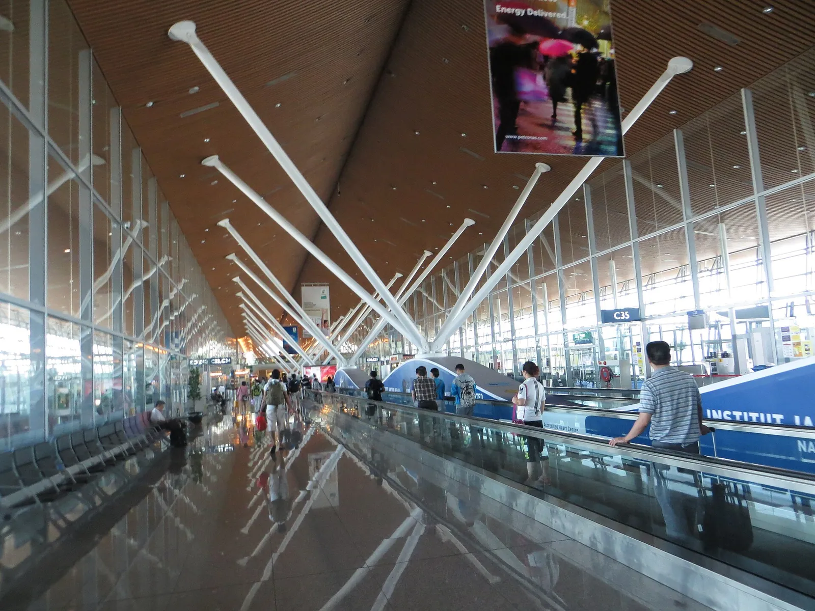 Passengers and a moving walkway beneath branching roof supports inside Kuala Lumpur International Airport