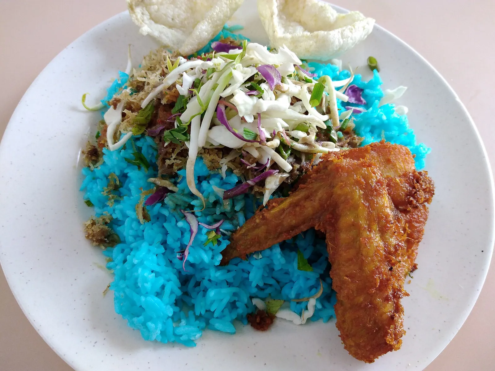 Blue nasi kerabu rice with shredded herbs and a fried chicken wing on a white plate