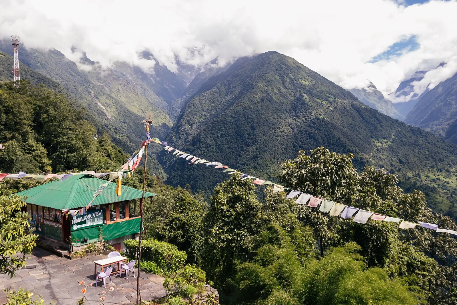 Pequeña terraza de un alojamiento con banderas de oración y colinas verdes en la ladera de Chhomrong
