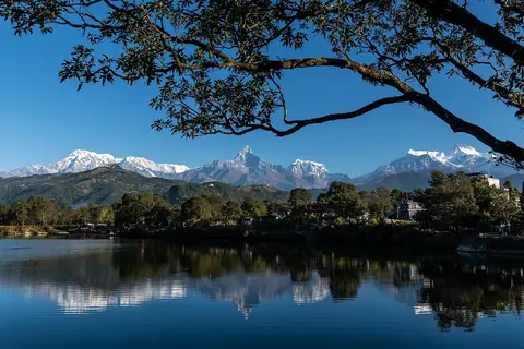 Machhapuchhre nevado y cumbres vecinas reflejados en las aguas tranquilas del lago Phewa, enmarcados por árboles