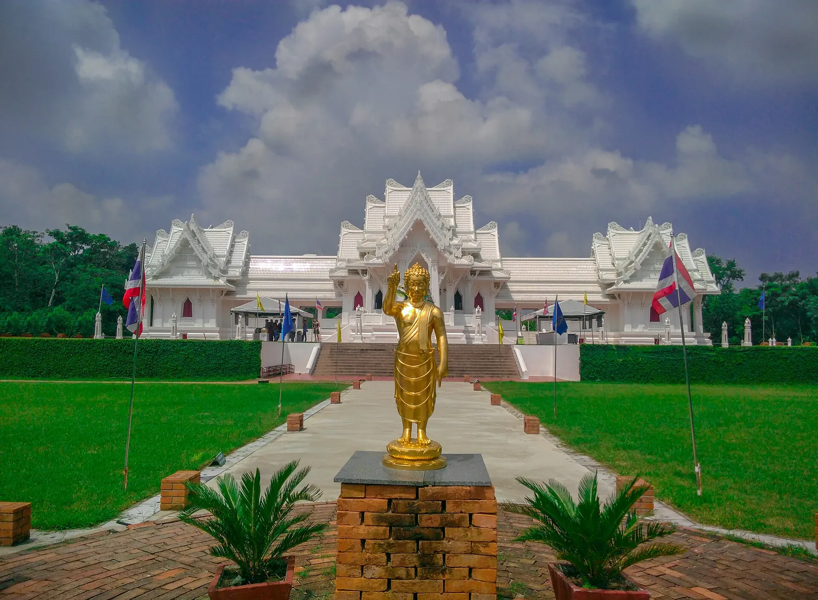 The white Royal Thai Monastery at Lumbini behind a lawn, a path and a golden Buddha statue