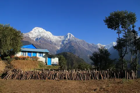 Una casa de tejado azul y una pila de leña en Ghandruk, con las cumbres nevadas del Annapurna al fondo bajo un cielo azul intenso