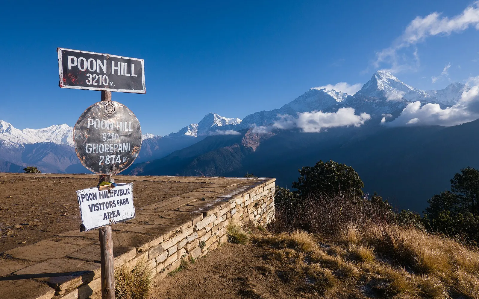 Ukazatel Poon Hill s výškou 3 210 m vedle vyhlídkové plošiny s kamennou zídkou a zasněženými vrcholy Annapurny v pozadí