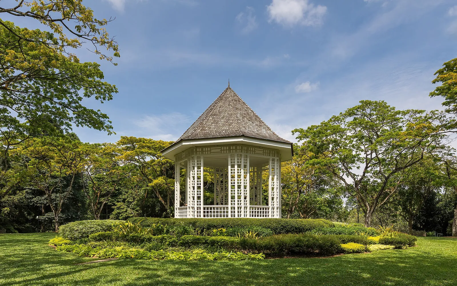 Hvid musikpavillon omgivet af grønne træer og solbeskinnet græs i Singapore Botanic Gardens