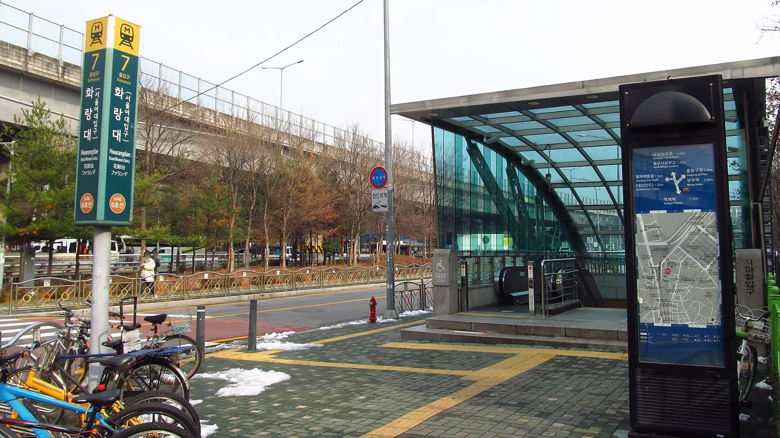 Exit 7 of Hwarangdae Station in Seoul, a glass-roofed subway entrance beside a tree-lined road with bicycles parked in front