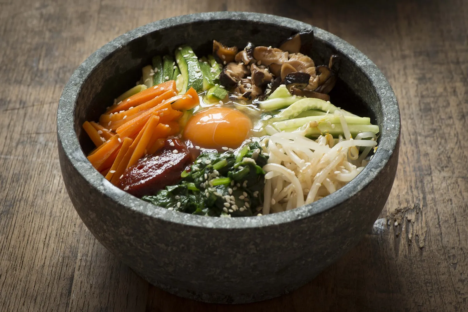 A hot stone bowl of dolsot bibimbap with rice topped with vegetables, spinach, bean sprouts and an egg yolk