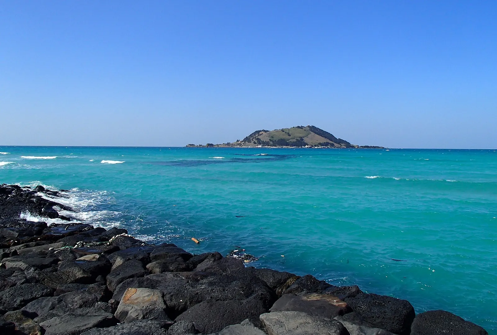 Turquoise sea breaking on black basalt rocks at Hyeopjae Beach, with the volcanic islet of Biyangdo on the horizon