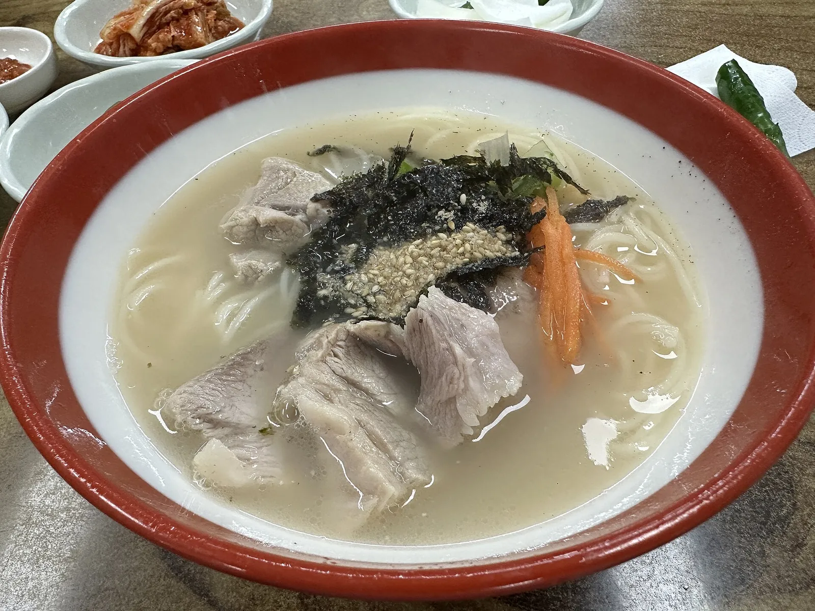 Bowl of gogi guksu, pork noodle soup topped with boiled pork slices, seaweed flakes and carrot, with side dishes
