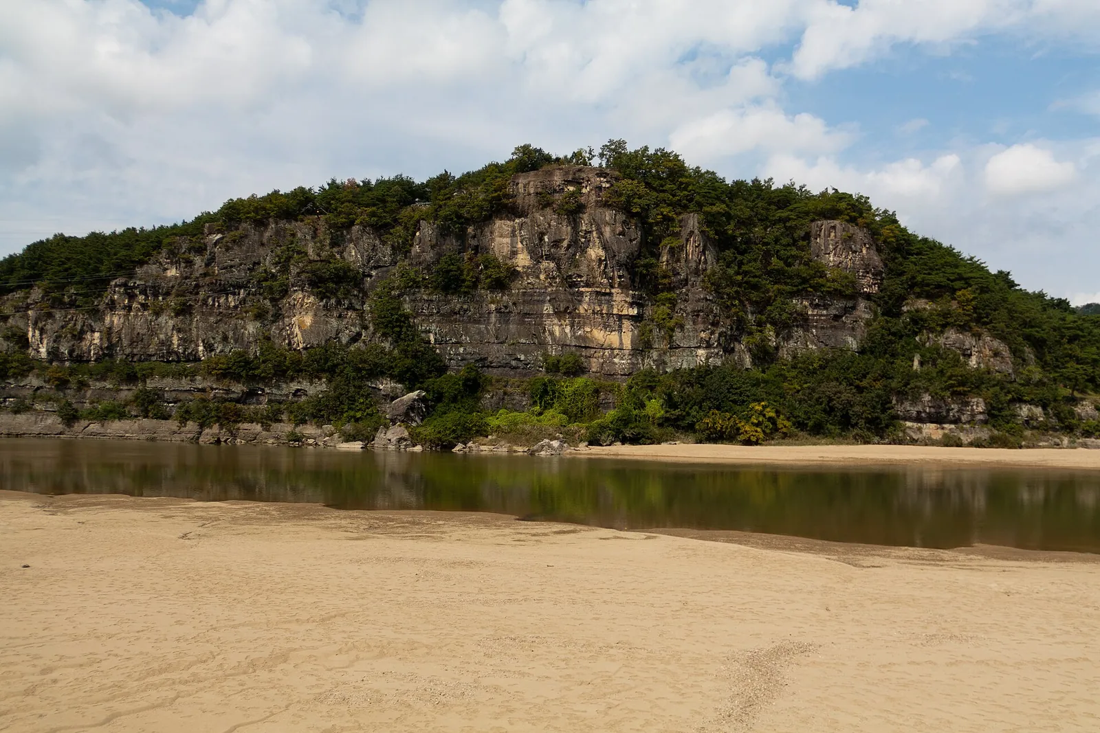 Buyongdae, a forested rock cliff rising behind a sandy riverbank of the Nakdonggang