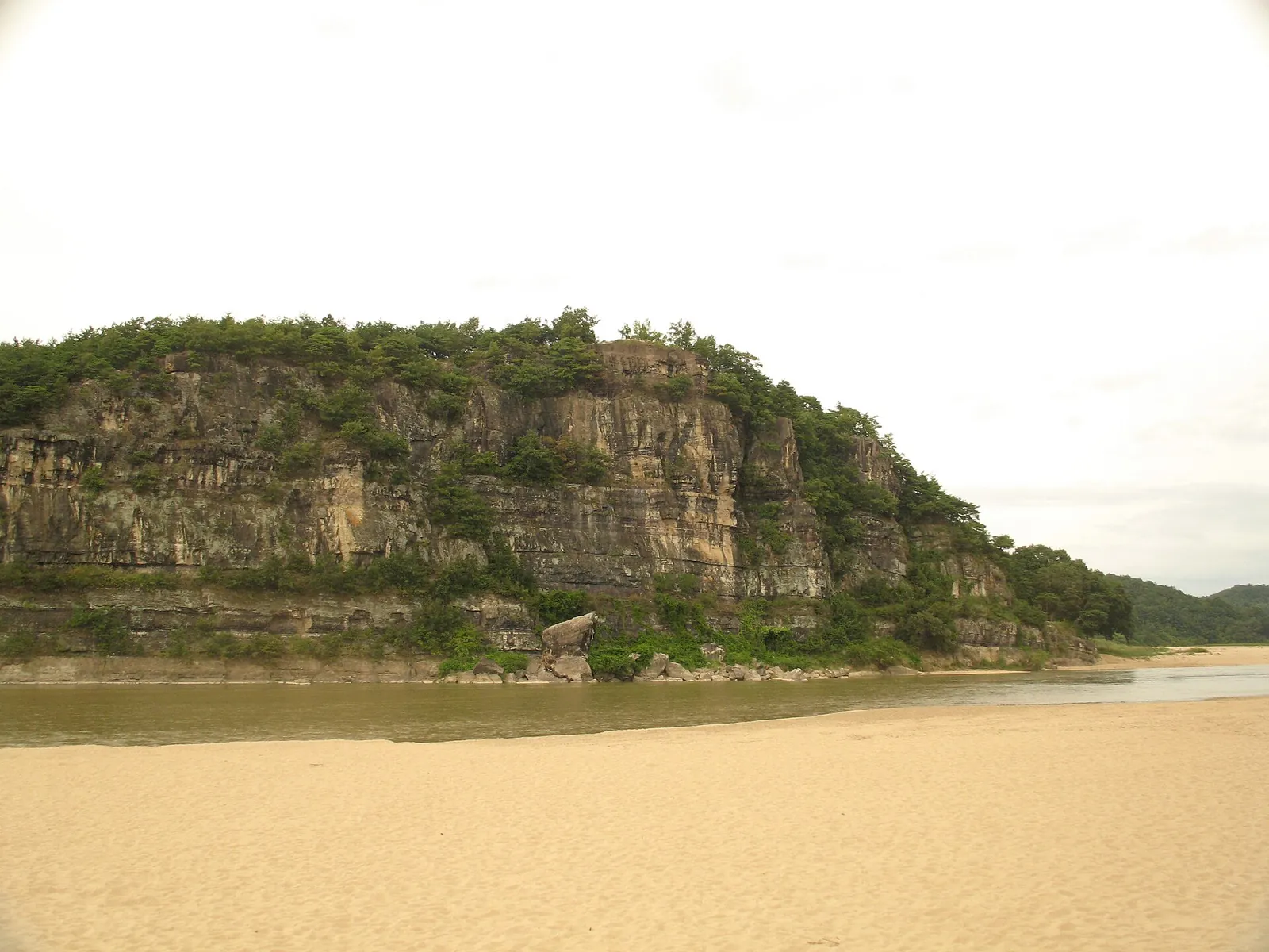 A wooded rock cliff above a sandy bank of the Nakdonggang River at Buyongdae