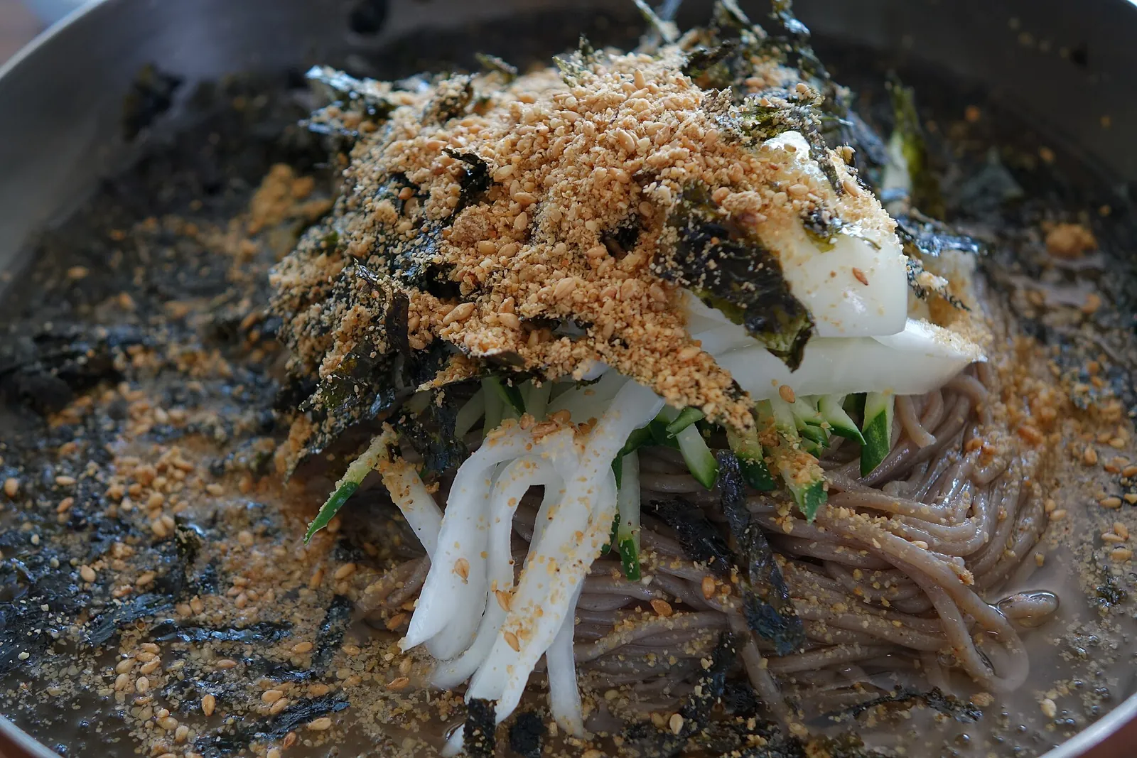 A bowl of makguksu buckwheat noodles topped with radish, cucumber, seaweed flakes and sesame