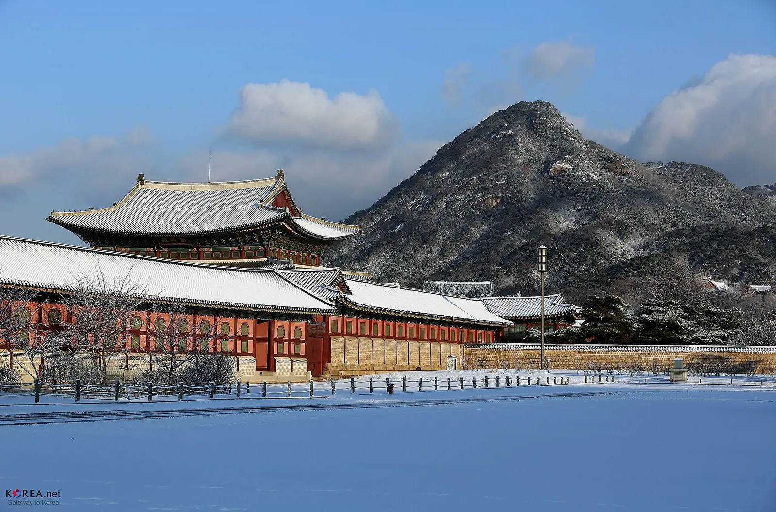 Gyeongbokgung Palace buildings and a snow-covered courtyard with a mountain behind under blue sky