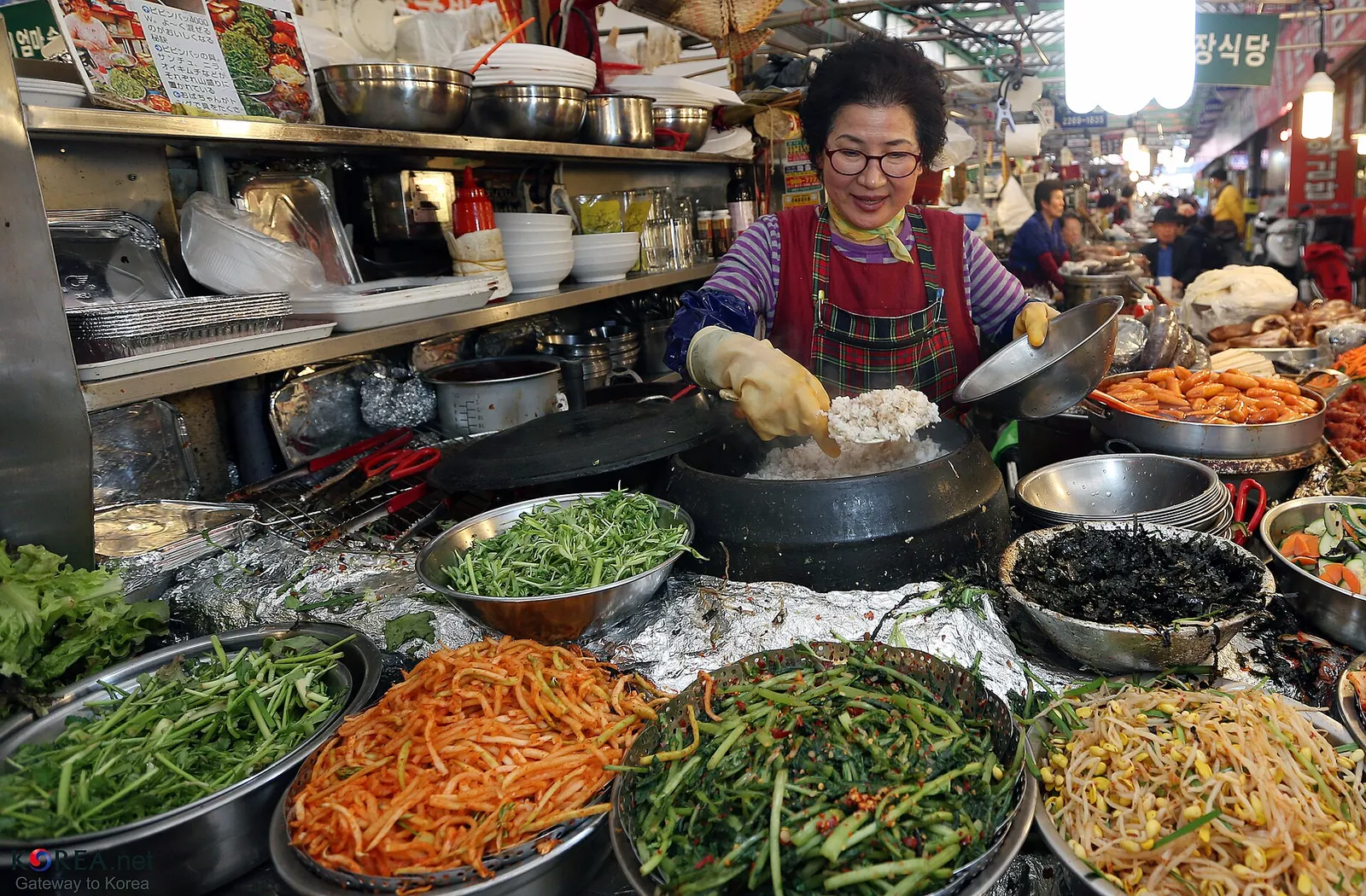 A vendor in an apron preparing bowls of seasoned vegetables for bibimbap at a Gwangjang Market food stall