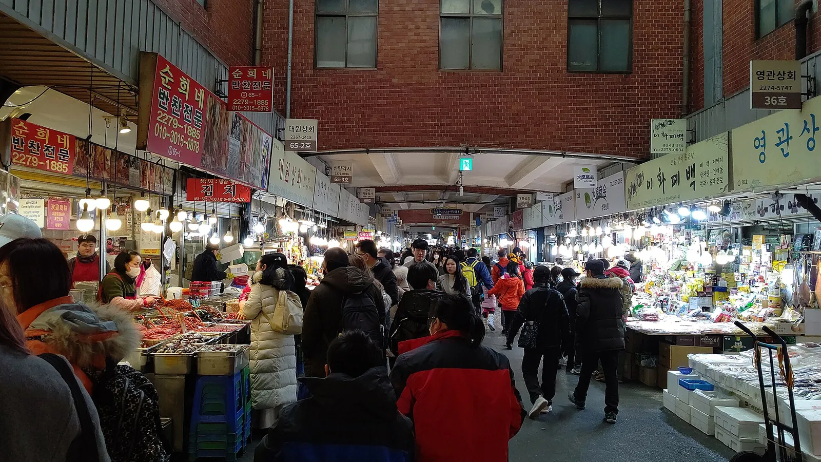 Shoppers walking down a crowded covered aisle lined with food and produce stalls in Gwangjang Market