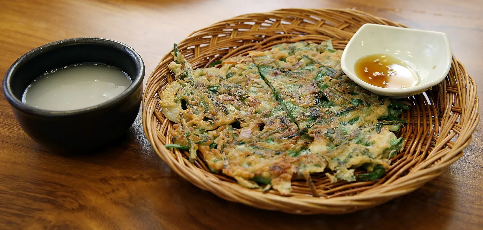 Savoury Korean pancake on a woven basket tray with a bowl of milky makgeolli and dipping sauce