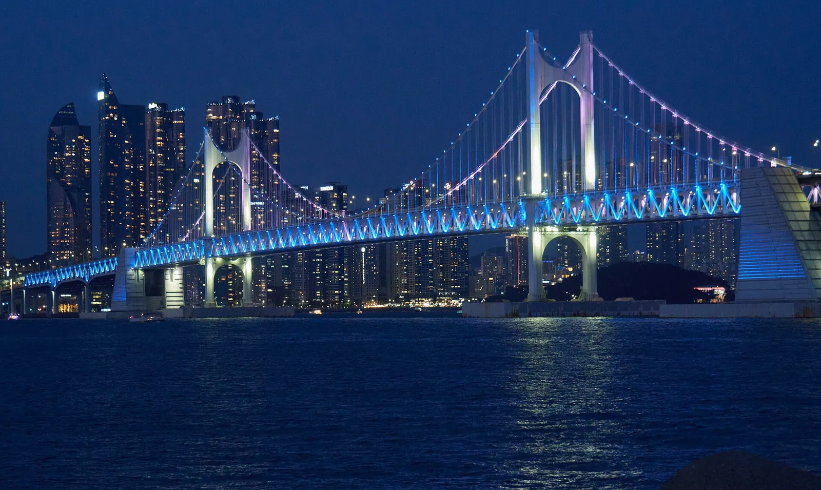 Gwangandaegyo Bridge lit up in blue at dusk, seen across the water from Gwangalli Beach in Busan