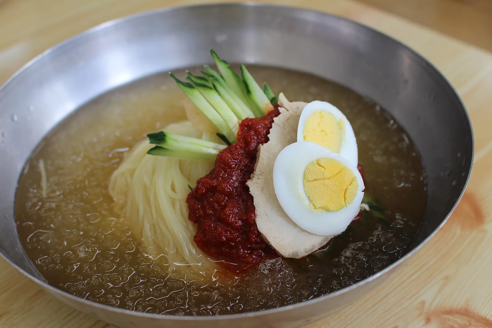 A metal bowl of milmyeon, cold wheat noodles in an icy broth topped with red pepper paste, sliced meat, cucumber and a halved egg