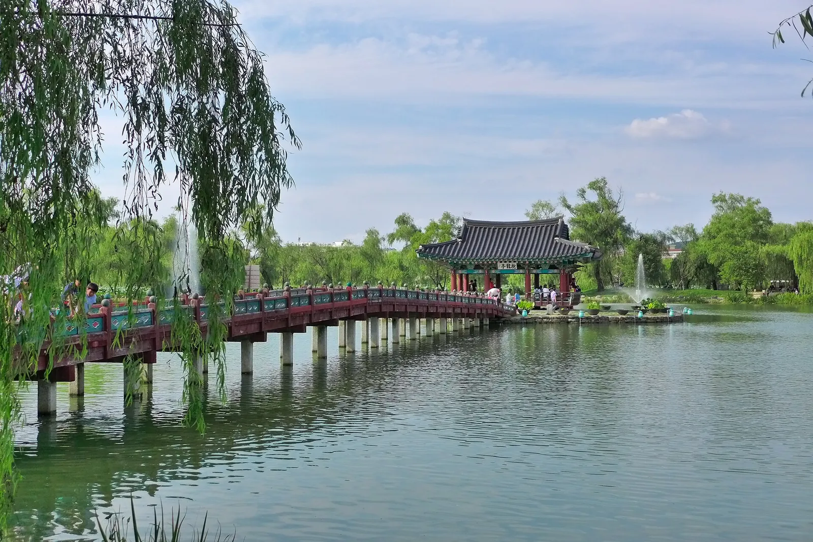 Red wooden bridge leading across Gungnamji Pond to a pavilion on an island, with willows on the bank