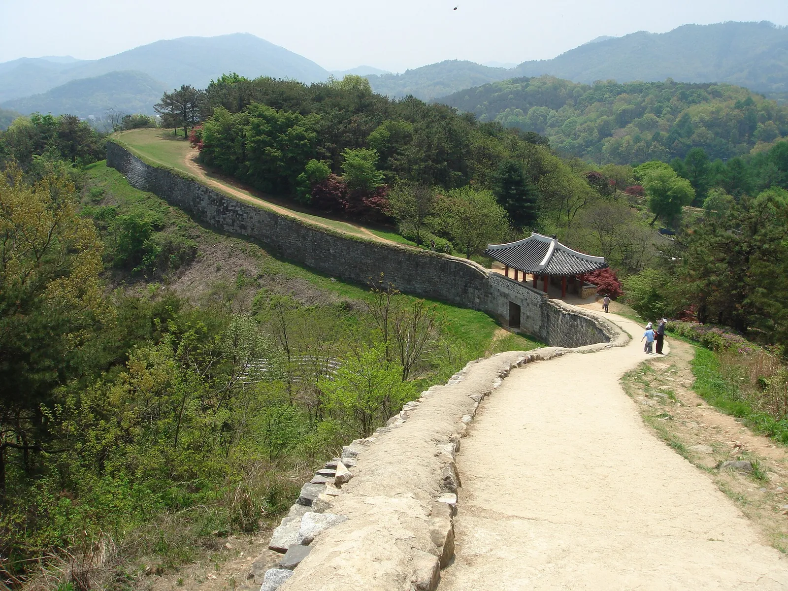 The stone wall of Sangdangsanseong Fortress running along a wooded ridge in Cheongju, with a walking path beside it and a pavilion below
