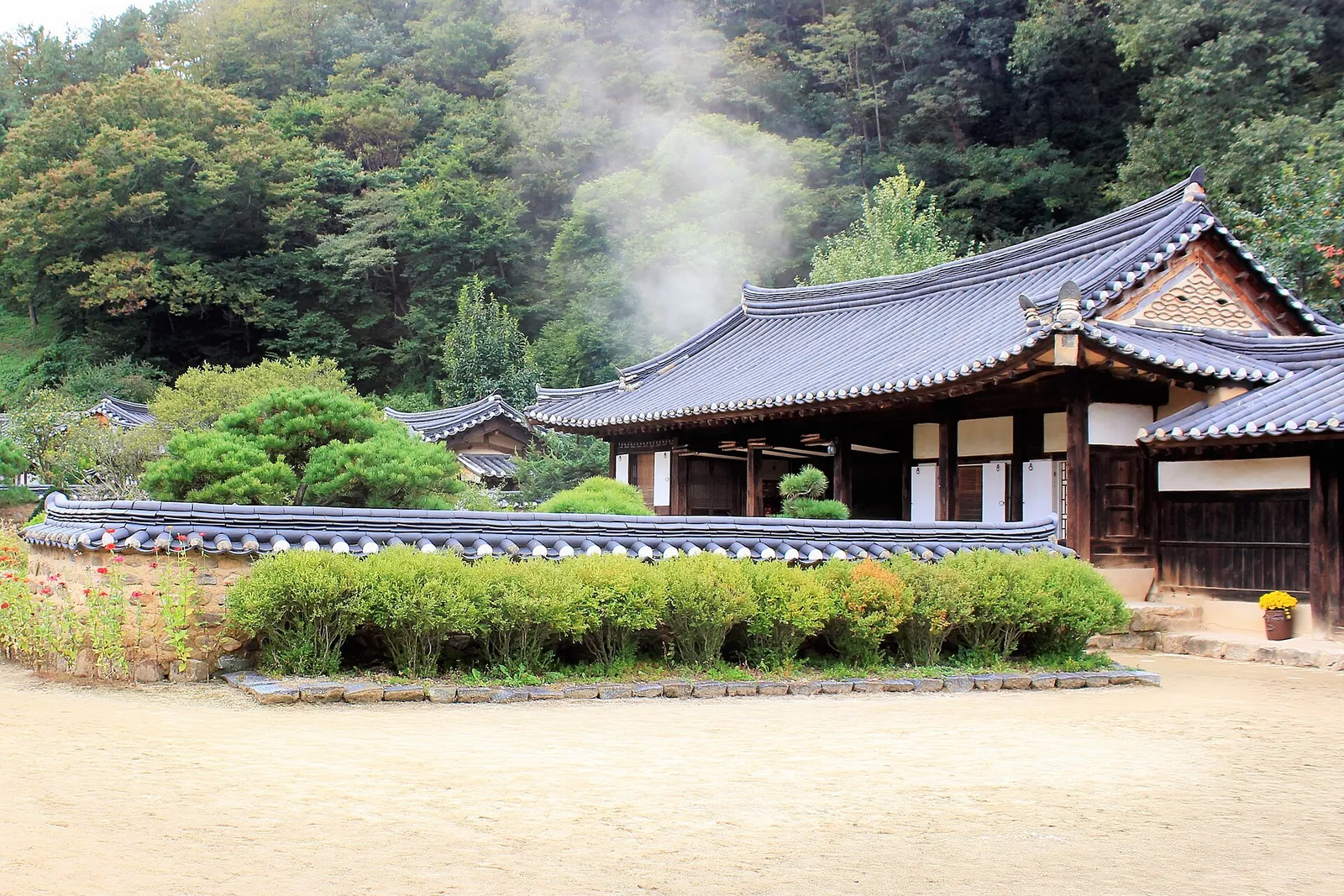Tiled-roof hanok buildings of Songso House behind a hedge and an open earthen yard, with smoke rising from the chimney