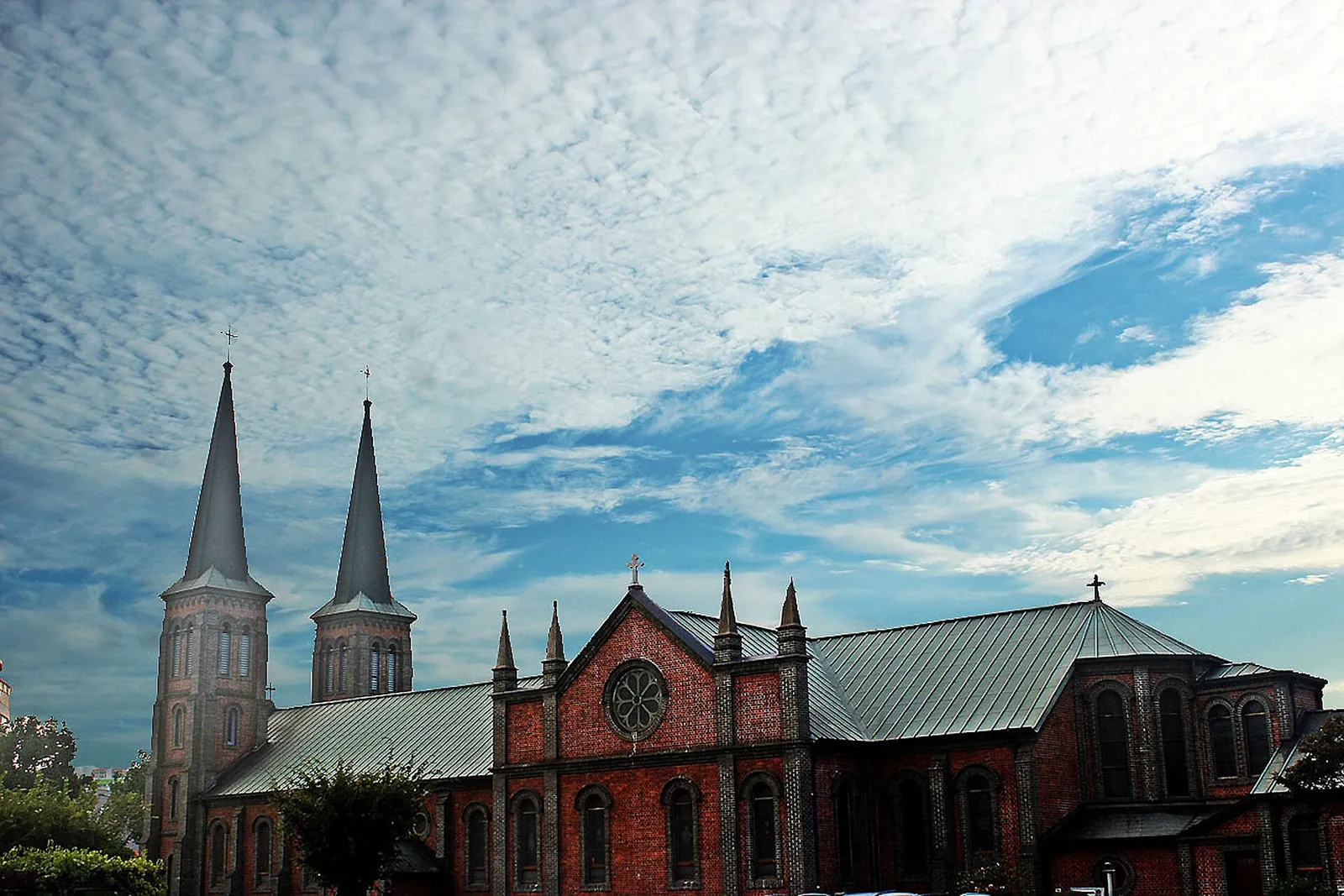 The red-brick Gyesan Cathedral in Daegu with its twin spires and green roofs under a blue sky with scattered clouds