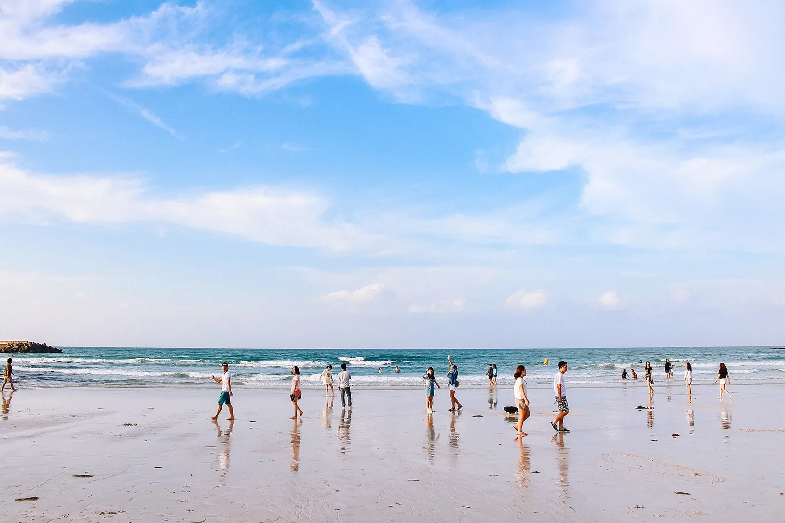 Visitors walking across wet white sand at the water's edge on Hyeopjae Beach, Jeju, under a blue sky