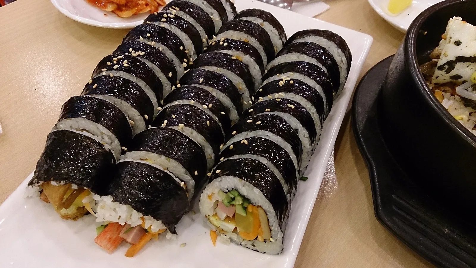 Rows of sliced gimbap on a white plate, with a stone bowl and side dishes beside it