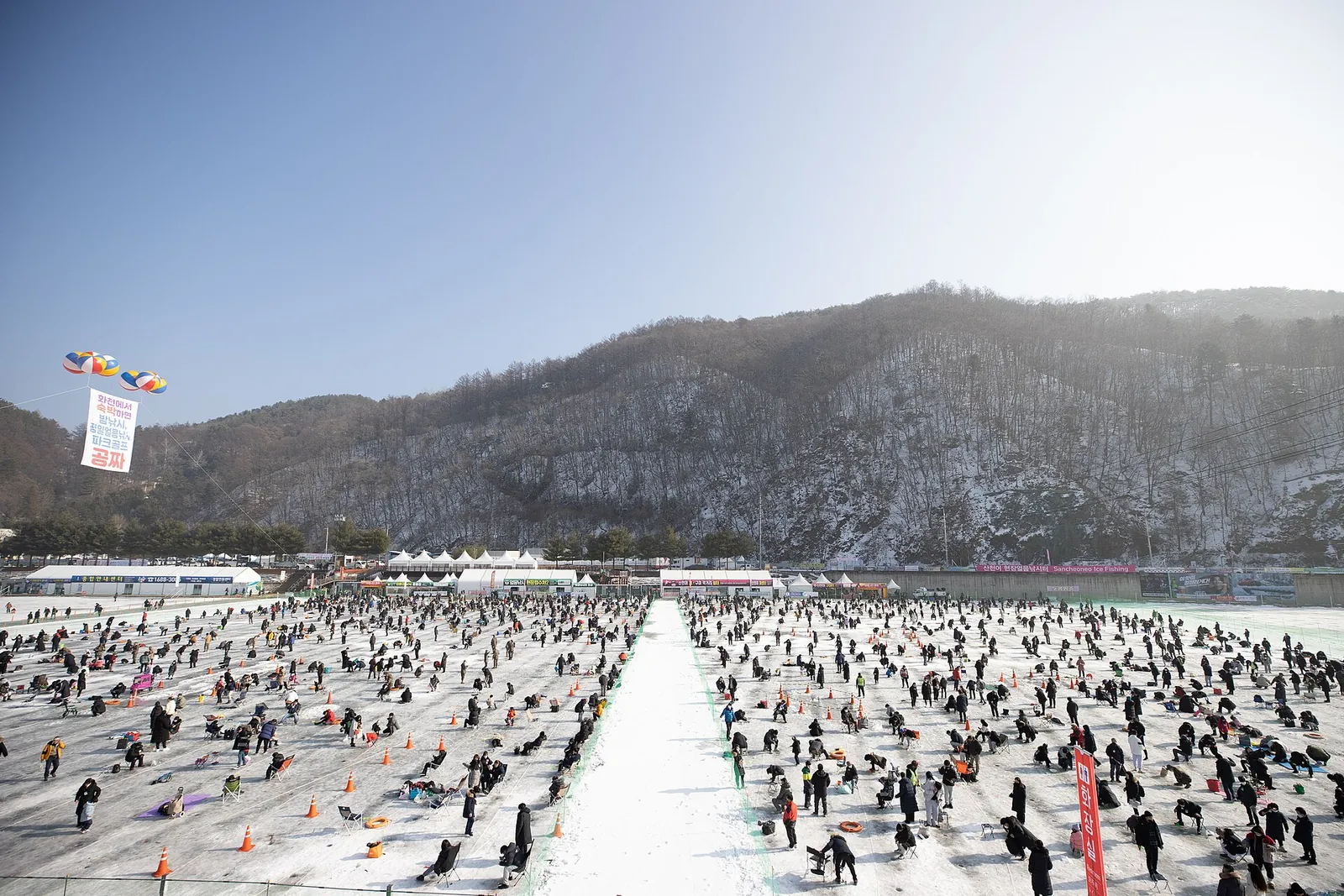 Hundreds of people ice fishing on a frozen river below a snowy hillside in Hwacheon
