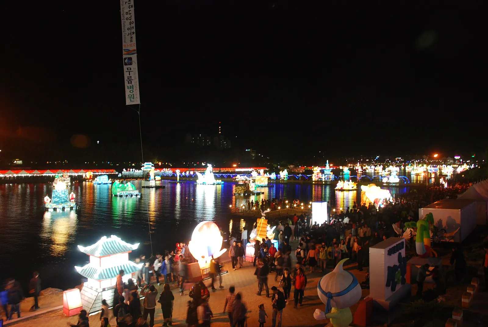 Illuminated lantern figures floating on the Namgang River at night, with crowds on the riverbank during the Jinju Namgang Yudeung Festival