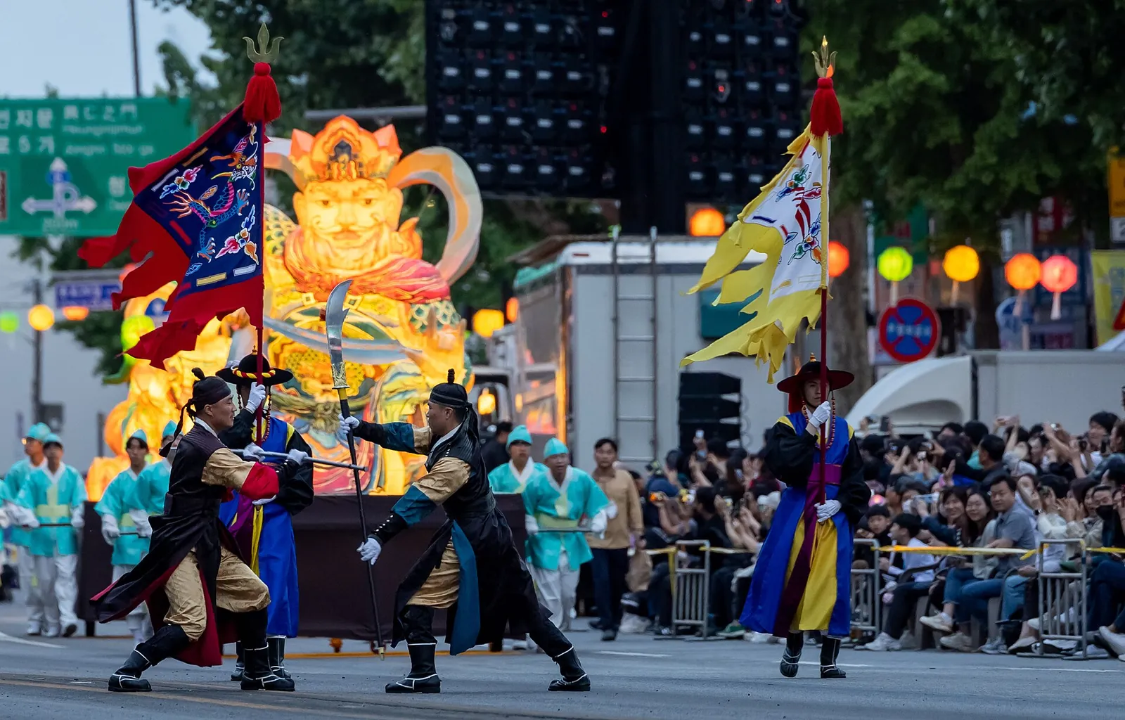 Performers in traditional costume and a large lantern float in a street parade lined with spectators during the Lotus Lantern Festival in Seoul