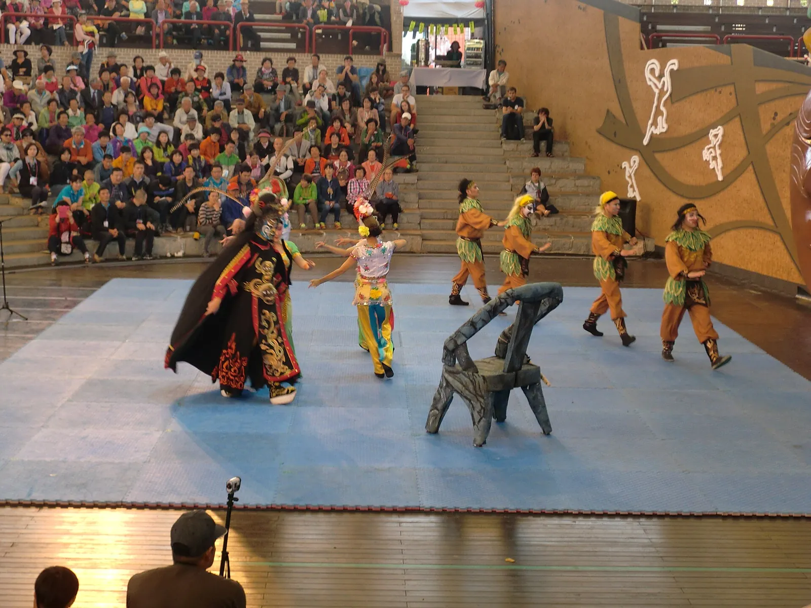 Masked dancers in colourful costumes performing on an open-air stage in front of a packed audience at the Andong Mask Dance Festival