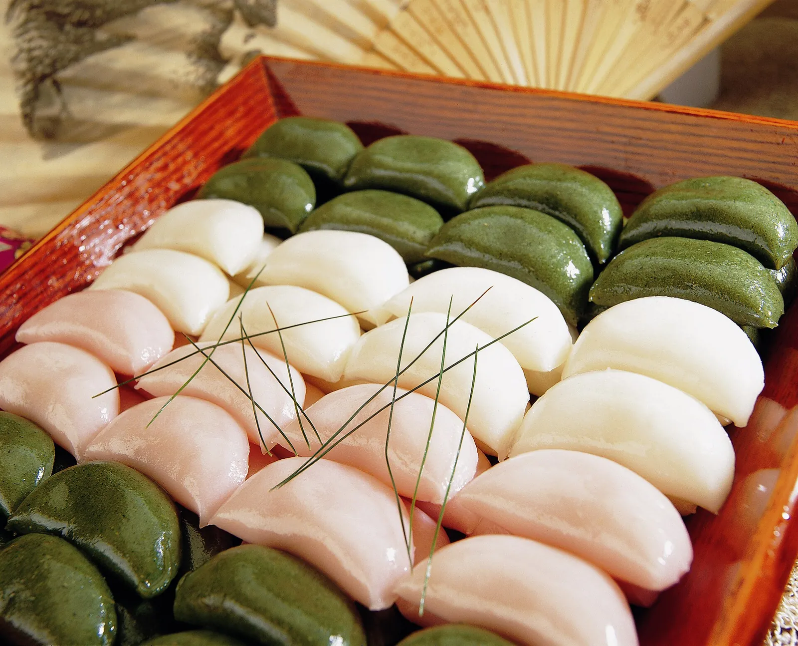 A wooden tray of songpyeon, half-moon rice cakes in white, pink and green, garnished with pine needles
