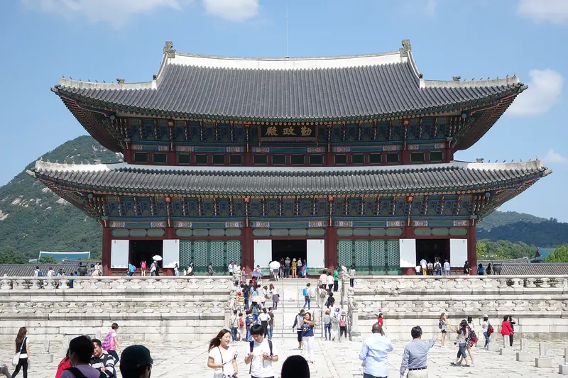 Geunjeongjeon, the two-tiered throne hall of Gyeongbokgung Palace in Seoul, under a blue sky with visitors on the stone courtyard
