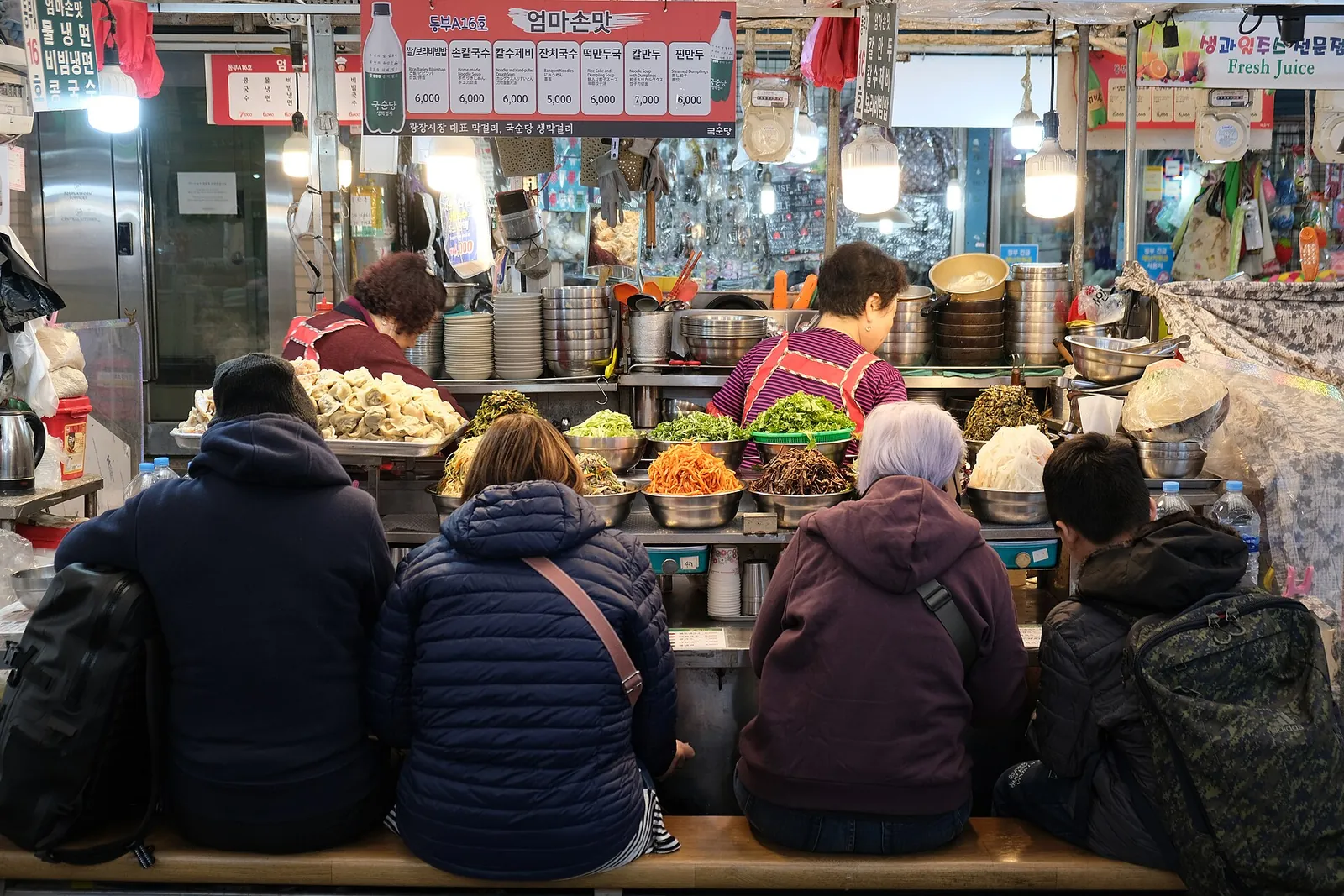 Visitors sitting on a bench at a Gwangjang Market food stall while vendors serve dishes from a counter piled with ingredients