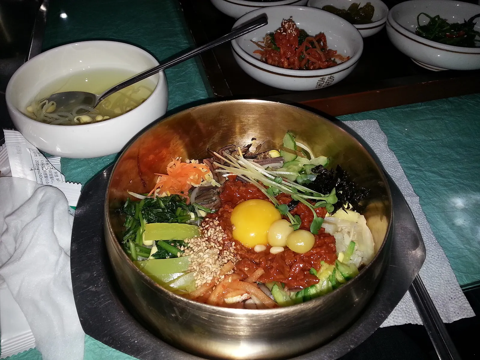 A brass bowl of Jeonju bibimbap with vegetables, a raw egg yolk and chilli paste, with bean sprout soup and side dishes