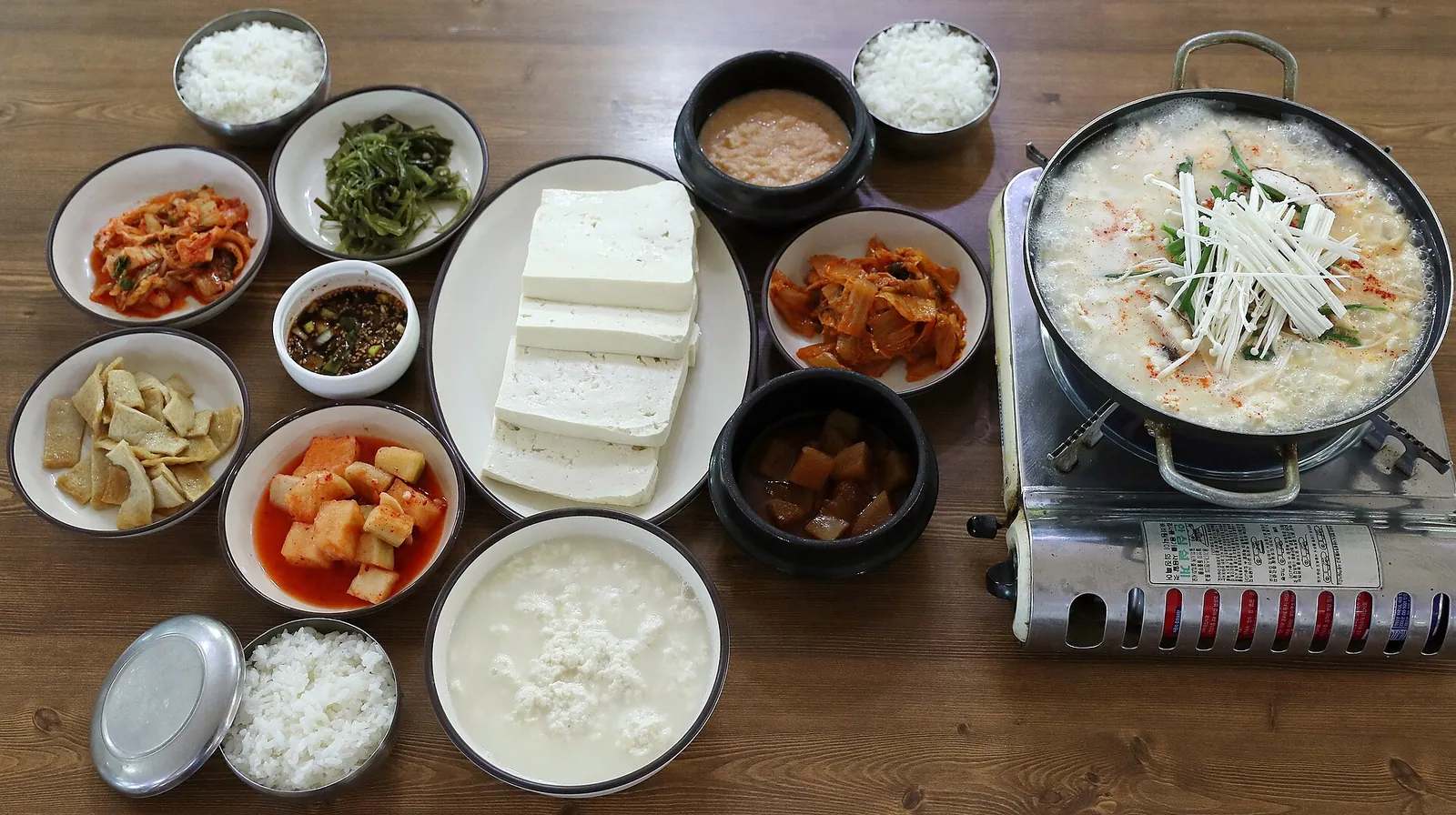 A Chodang sundubu meal: a pot of soft tofu stew on a burner, a plate of white tofu blocks and many side dishes