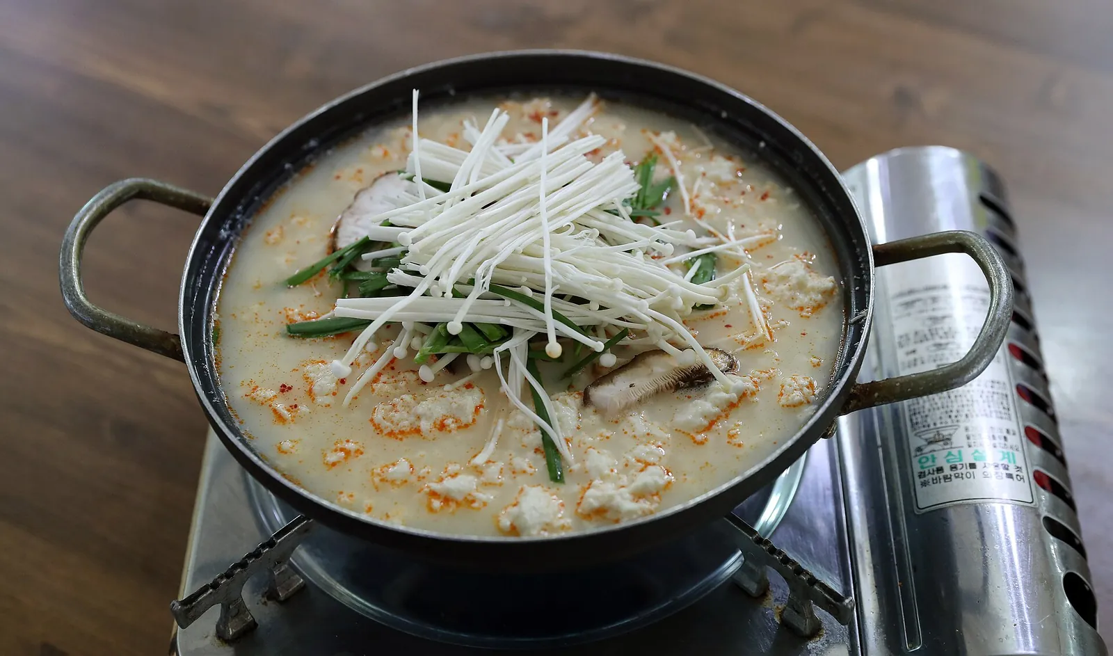 A pot of sundubu, soft tofu stew topped with enoki mushrooms and green onion, on a tabletop gas burner