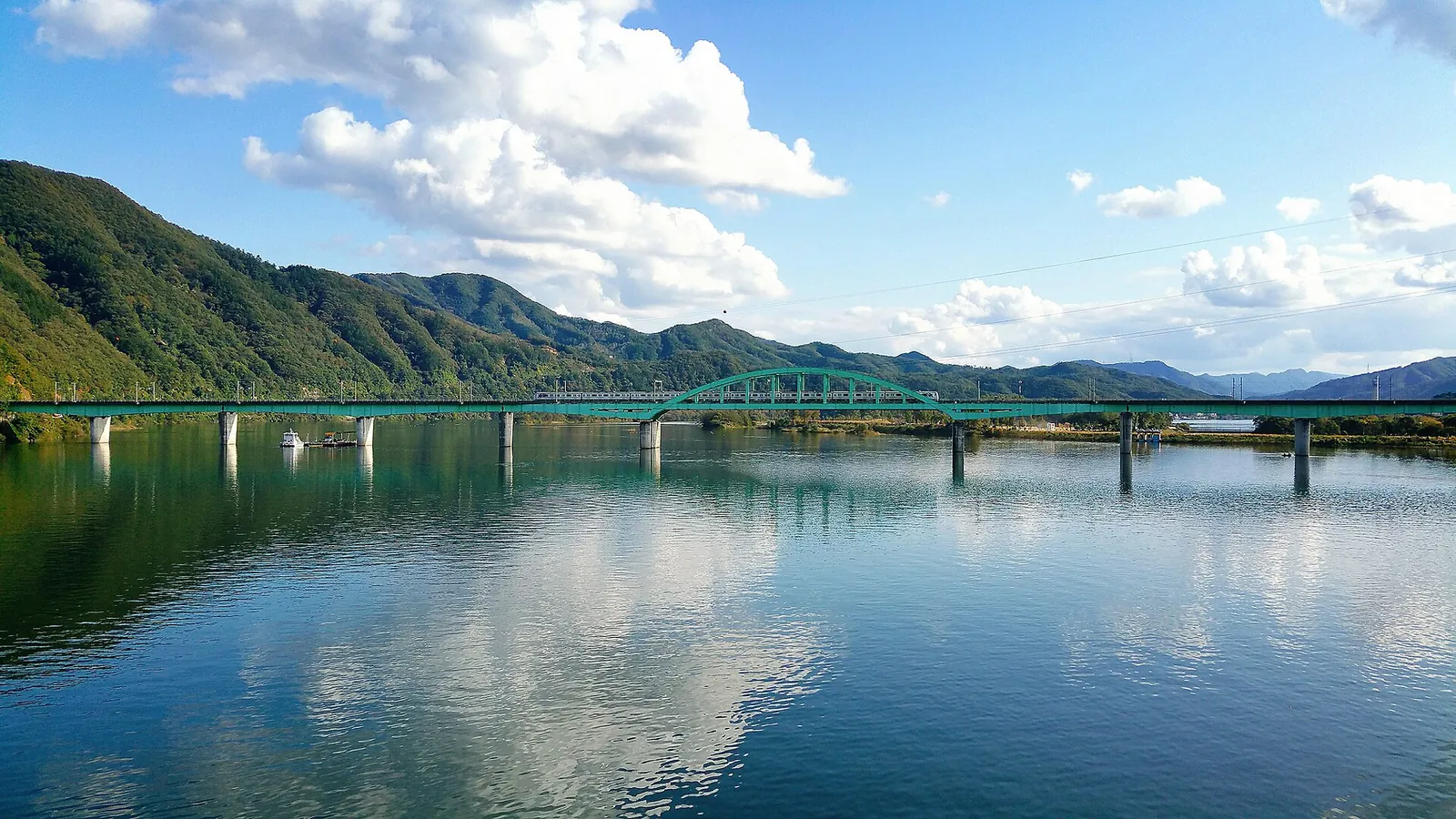 Green arched railway bridge crossing a calm, wide river framed by mountains
