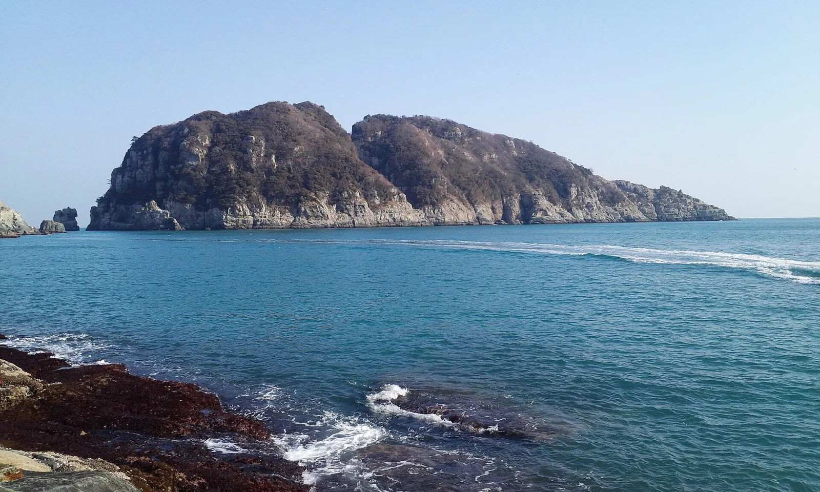 A steep rocky islet rising from blue water, with surf breaking over dark rocks in the foreground